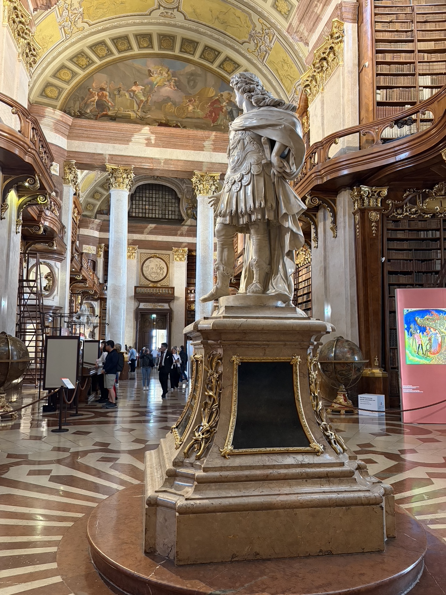 A perfect addition to your Vienna bucket list, the ornate State Hall of the Austrian National Library in Vienna, with floor-to-ceiling frescoes, towering bookshelves, and baroque architecture